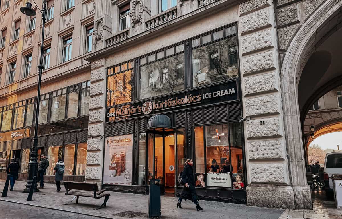 Exterior view of Café Molnár’s Kürtőskalács and Ice Cream shop. The storefront features large windows, a sign with the café’s name, and a poster showing their pastries. Pedestrians walk by on the sidewalk.