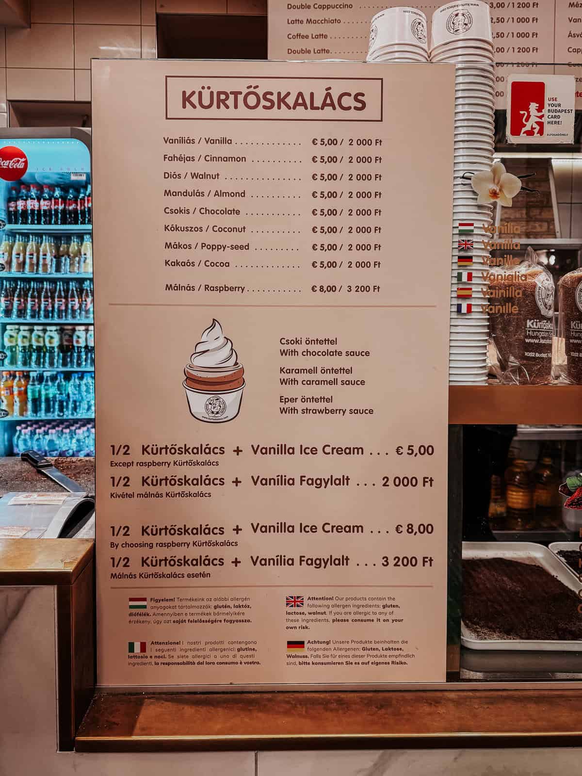 A menu board at Café Molnár’s displaying various flavors of Kürtőskalács, a Hungarian pastry. Flavors listed include vanilla, cinnamon, walnut, almond, chocolate, coconut, poppy-seed, cocoa, and raspberry. Prices are shown in euros and Hungarian forints. The board also mentions options to add vanilla ice cream with different sauces.