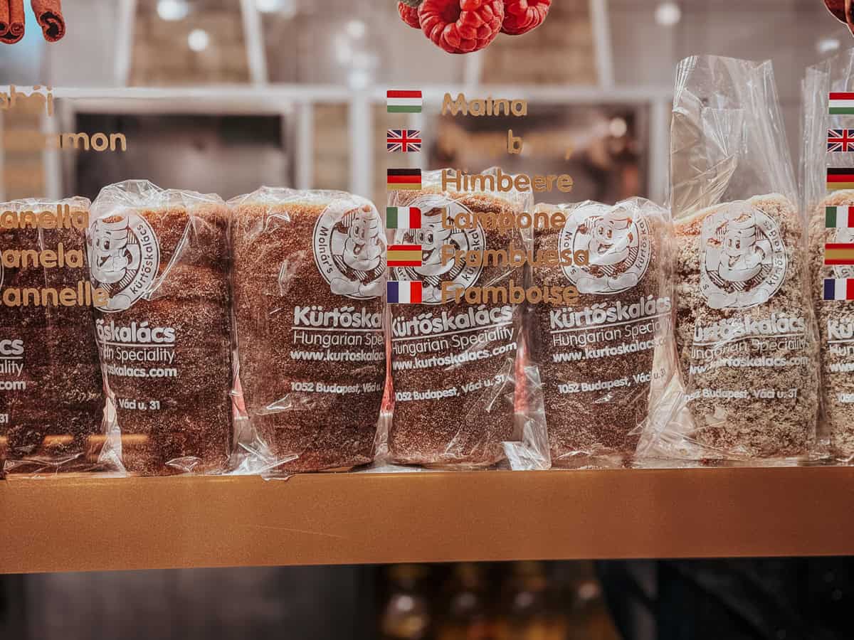 Several packaged Kürtőskalács pastries lined up on a shelf, each wrapped in clear plastic and labeled with the café's logo and details. The flavors are written in multiple languages on the glass above the pastries, including cinnamon, raspberry, and more.