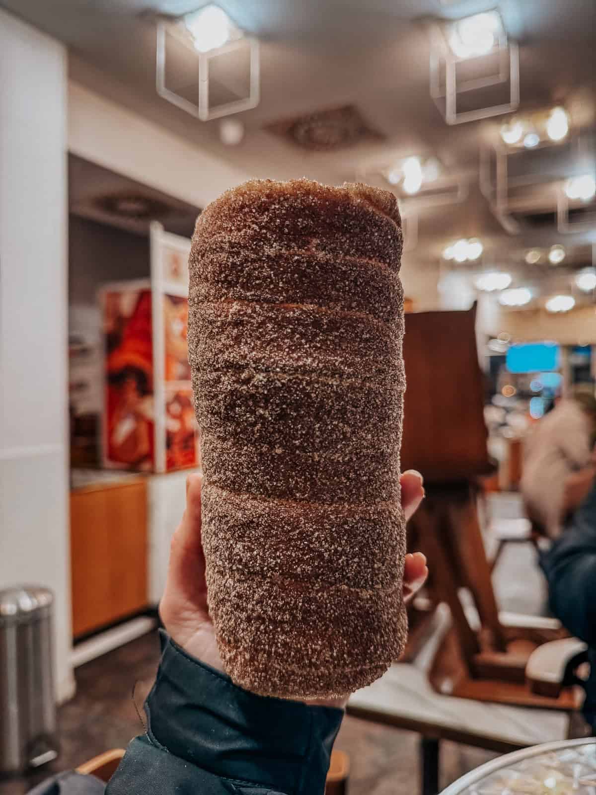 A hand holding a freshly baked Kürtőskalács, coated in sugar. The background shows the interior of Café Molnár’s, with tables and a warm, inviting atmosphere.