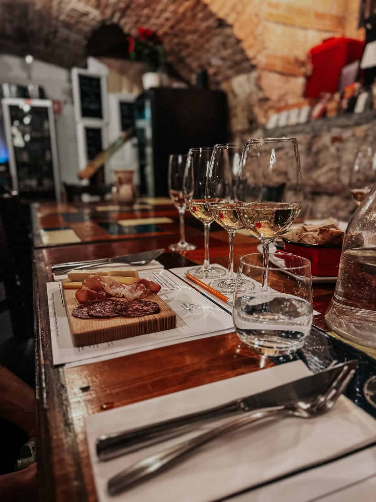 A rustic restaurant setting with a table featuring wine glasses, a charcuterie board with various meats and cheeses, and place settings. The ambiance is cozy with brick archways and warm lighting.