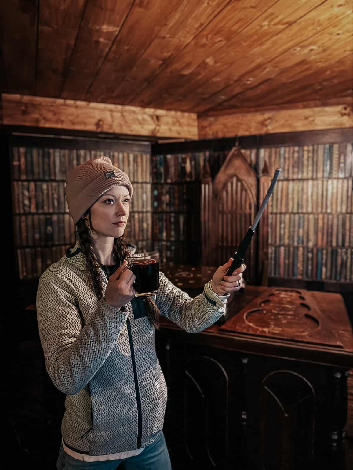 A woman dressed warmly, holding a drink and a wand, posing in a room that resembles a magical library with wooden accents.