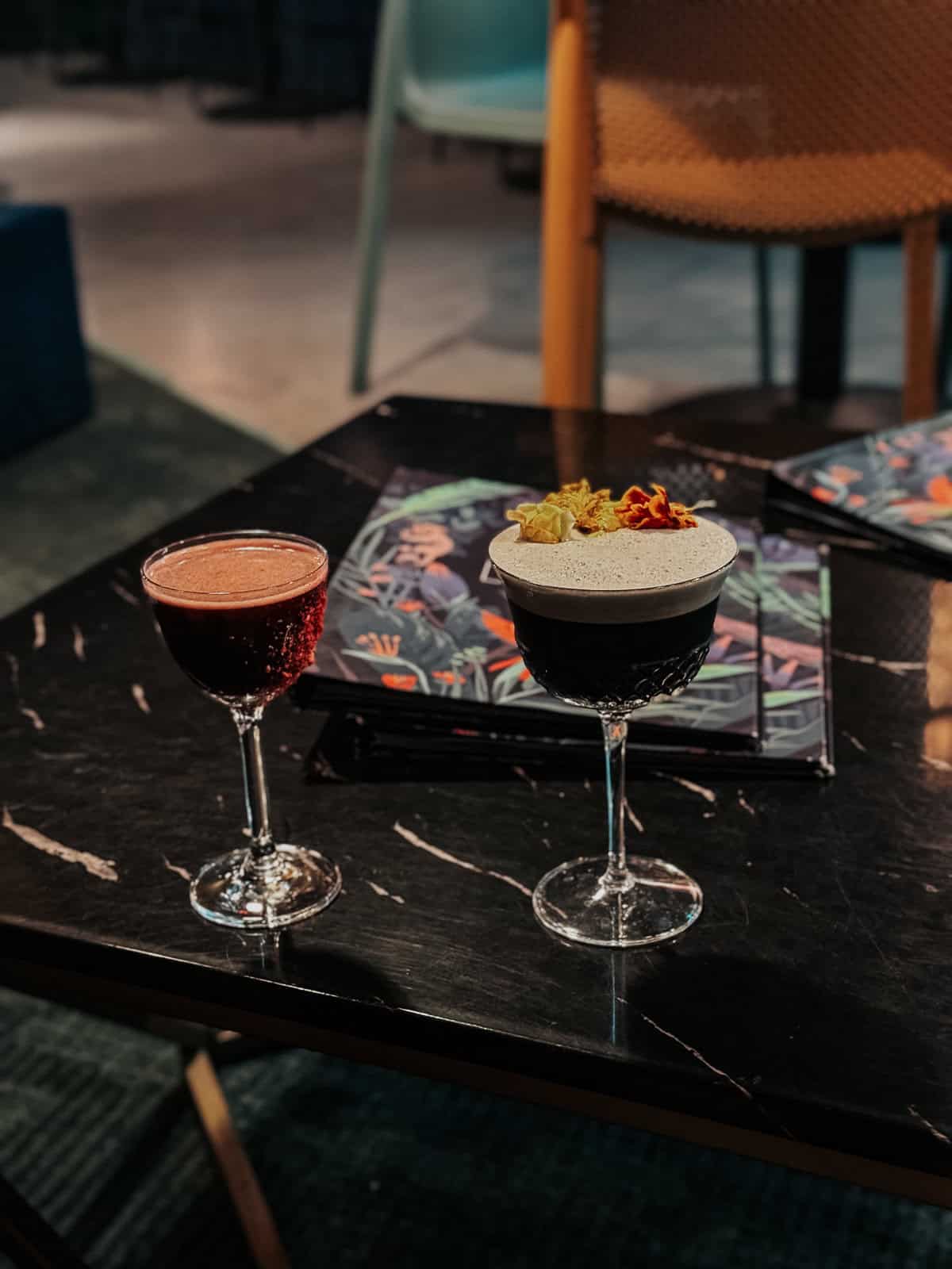 Two beautifully garnished cocktails on a dark marble table, set against a backdrop of modern, colorful decor.