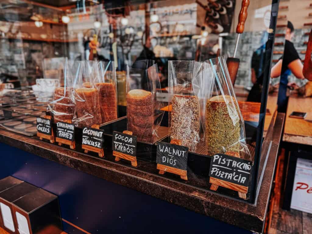 A display of various chimney cakes covered in different toppings, including walnut, cinnamon, coconut, cocoa, and pistachio. The cakes are neatly wrapped in plastic, and small chalkboard signs label each flavor.