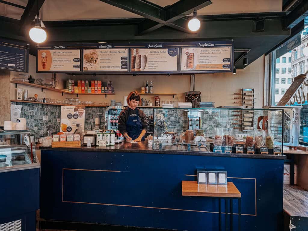 The interior of a chimney cake shop with a counter displaying different flavors of chimney cakes. A staff member is preparing a cake behind the counter, and the menu board above lists various chimney cake options and prices.