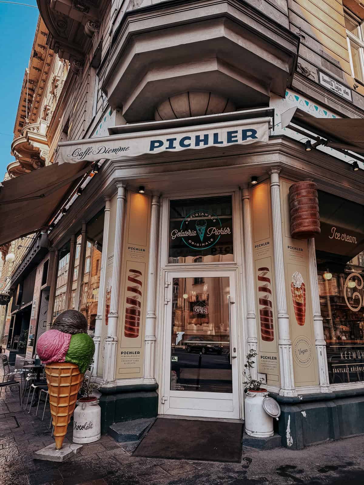 The exterior of Gelateria Pichler, a café and ice cream shop, with a large ice cream cone statue outside. The shop's entrance is decorated with images of chimney cakes and ice cream.