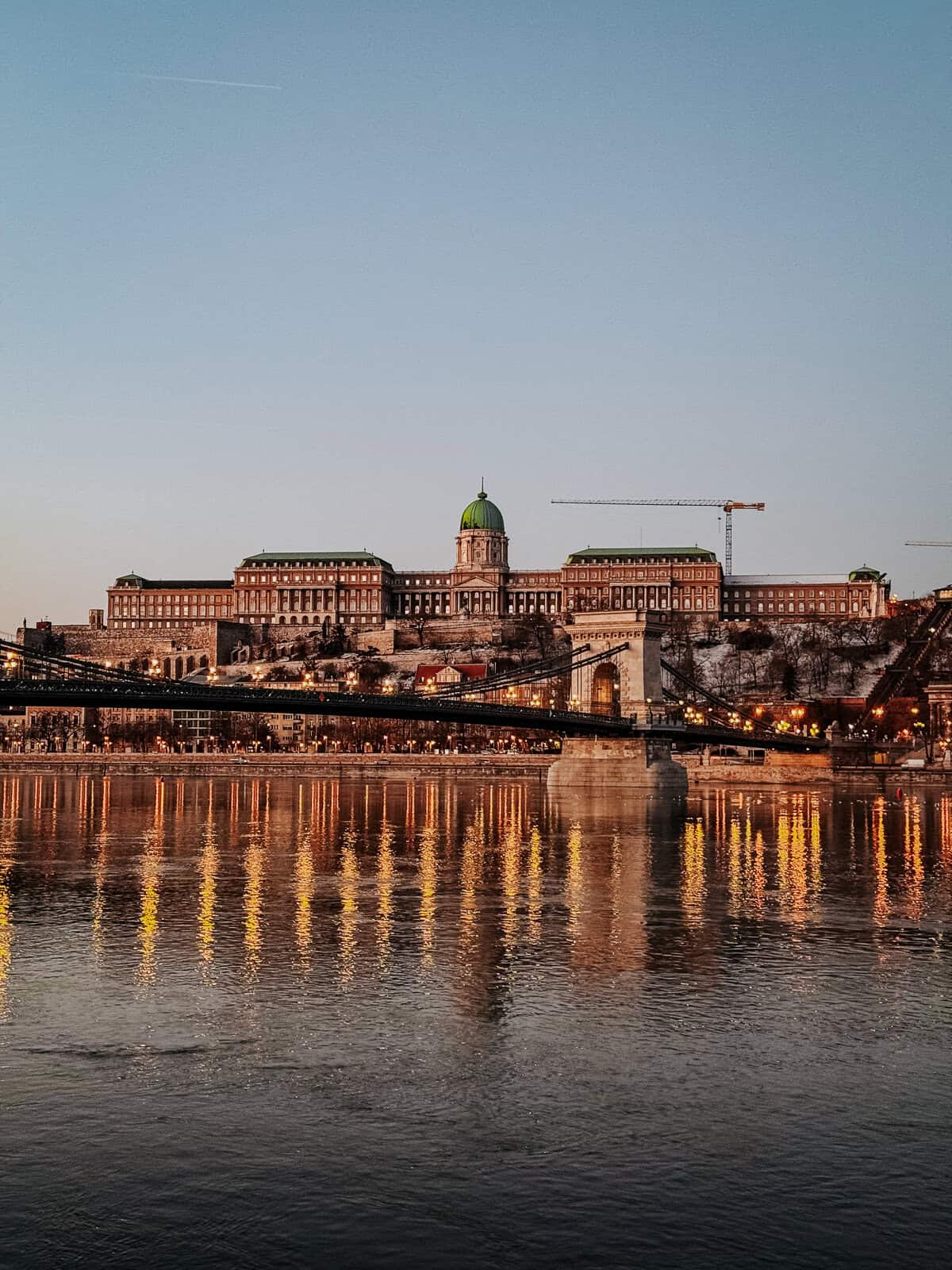 The Buda Castle in Budapest at dusk, with the Chain Bridge in the foreground, reflecting the warm lights on the Danube River.