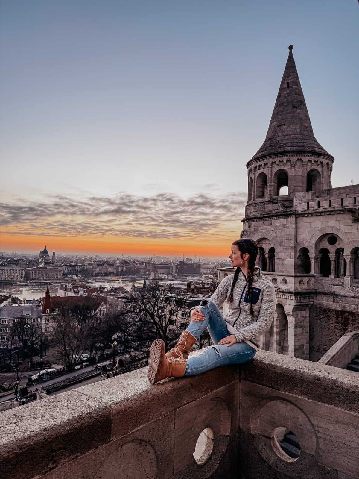 A woman sitting on the edge of Fisherman's Bastion in Budapest, enjoying a sunset view over the city and the Danube River