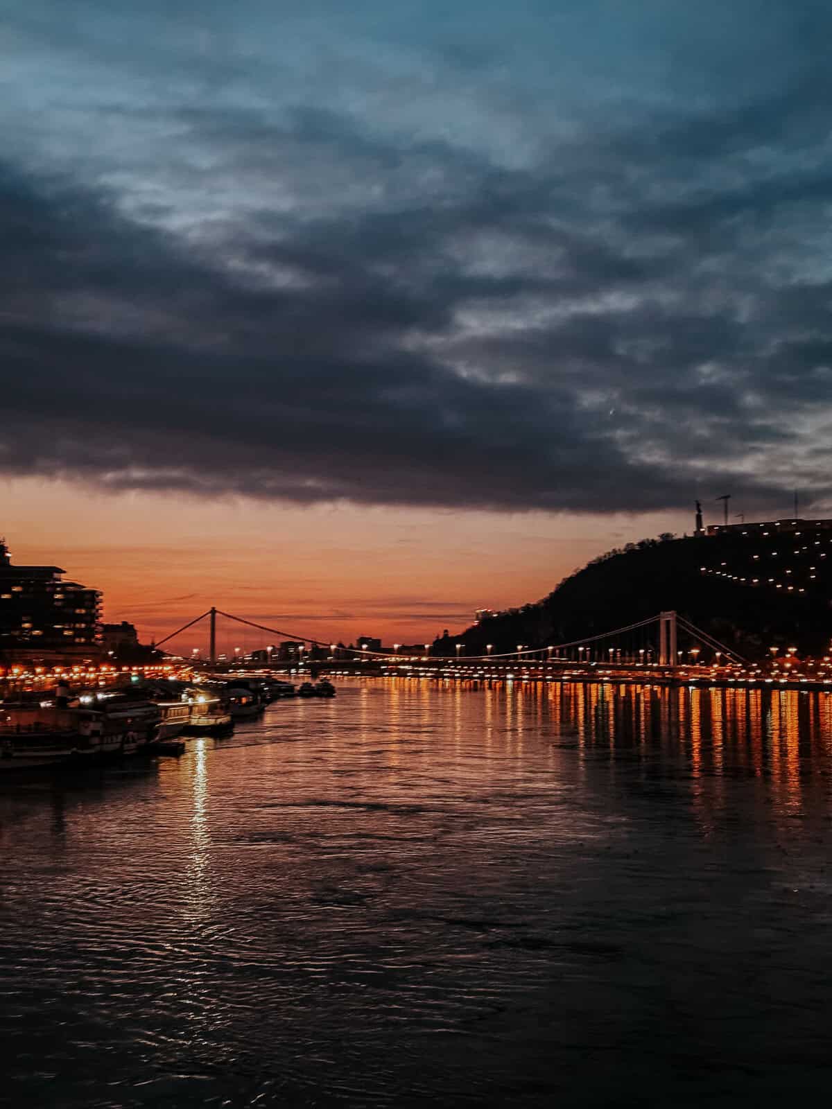 The Danube River at sunset with dark clouds overhead, city lights reflecting on the water, and a bridge in the distance.
