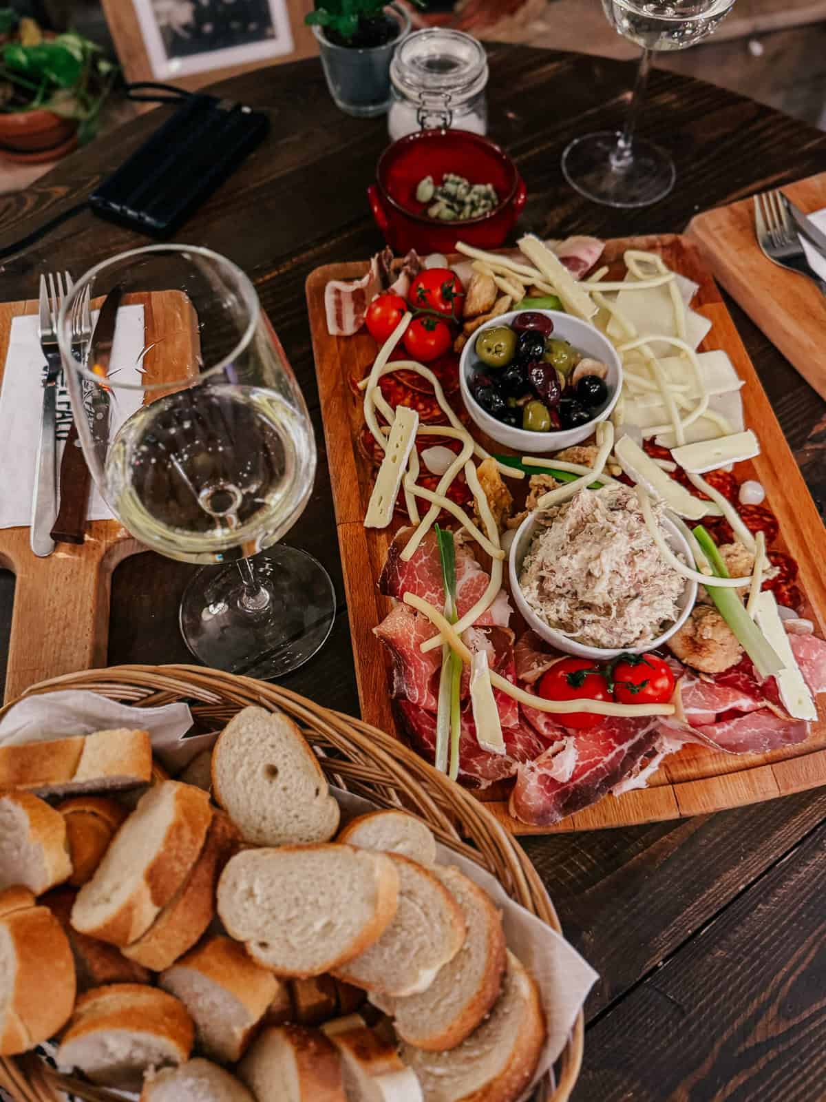 A charcuterie board with assorted meats, cheeses, olives, tomatoes, and bread, accompanied by glasses of white wine on a wooden table