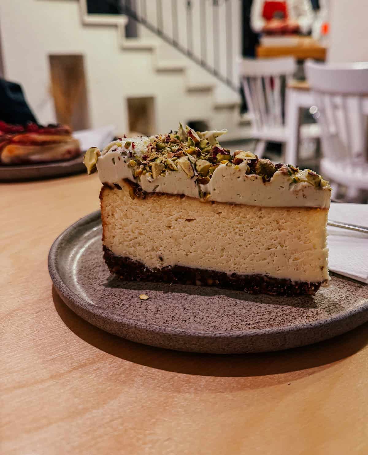 A slice of cheesecake topped with chopped pistachios served on a gray plate, with a staircase and dining area in the background.