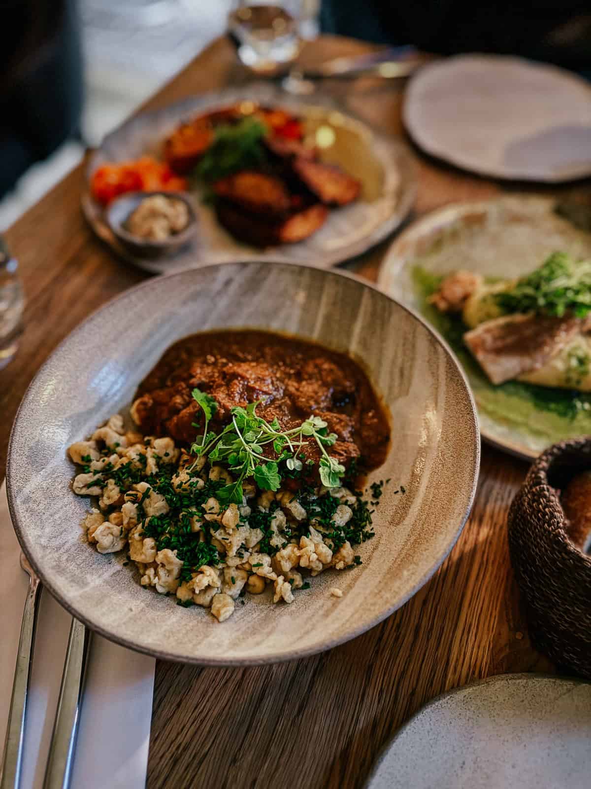 A plate of traditional Hungarian goulash with spaetzle, garnished with fresh herbs, served at a restaurant with other dishes in the background