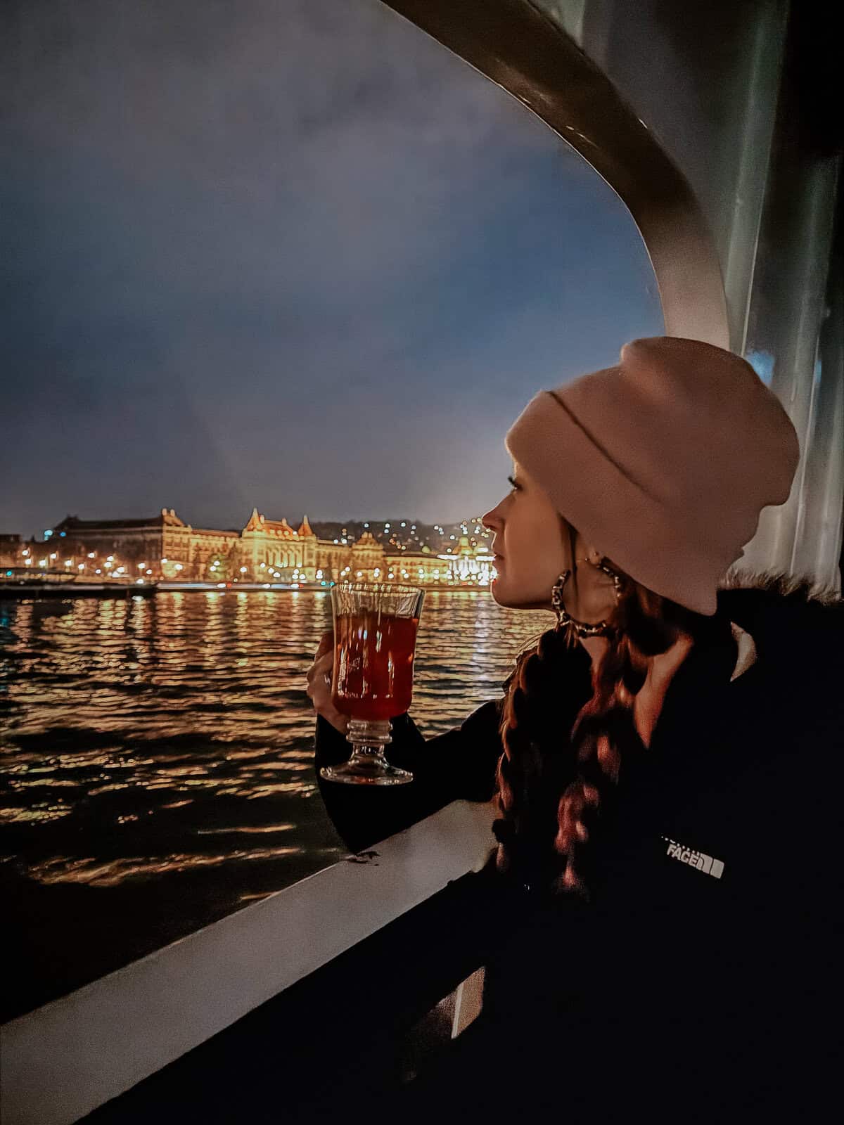 A woman in a pink hat enjoying a warm drink on a nighttime river cruise, with the illuminated Budapest skyline in the background