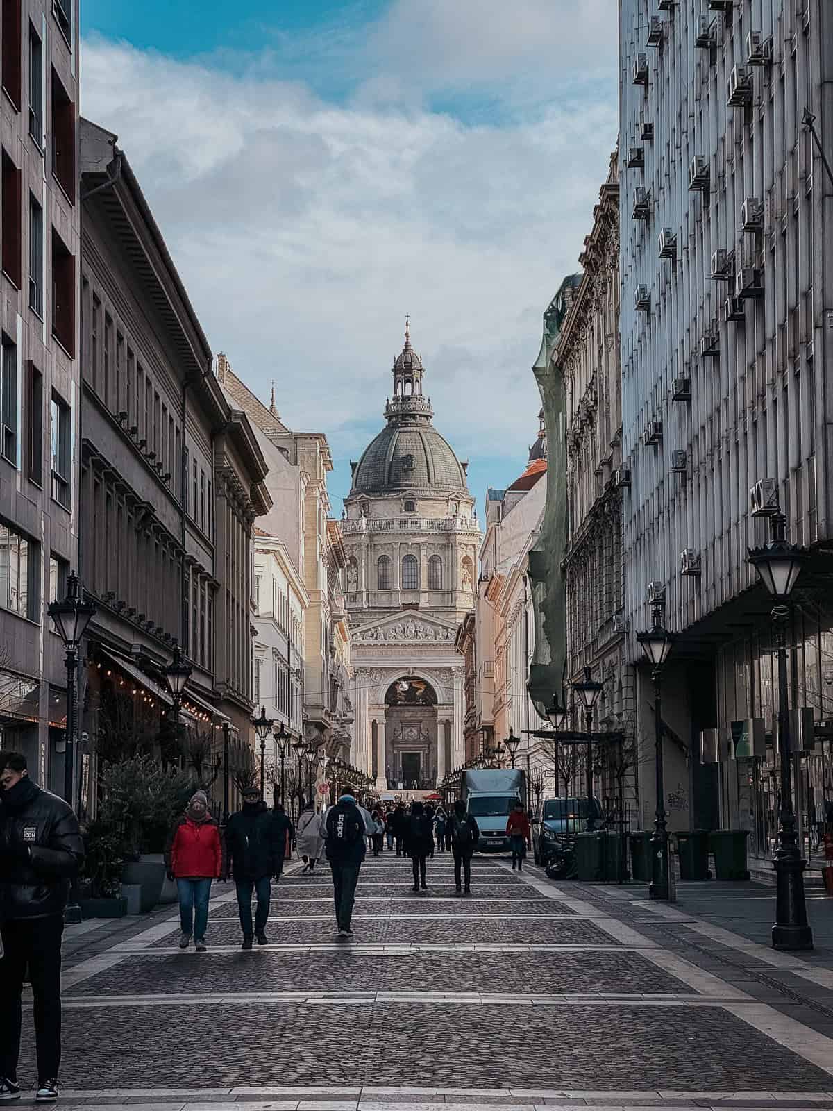 A bustling street in Budapest leading to St. Stephen's Basilica, lined with buildings and people walking, under a partly cloudy sky.