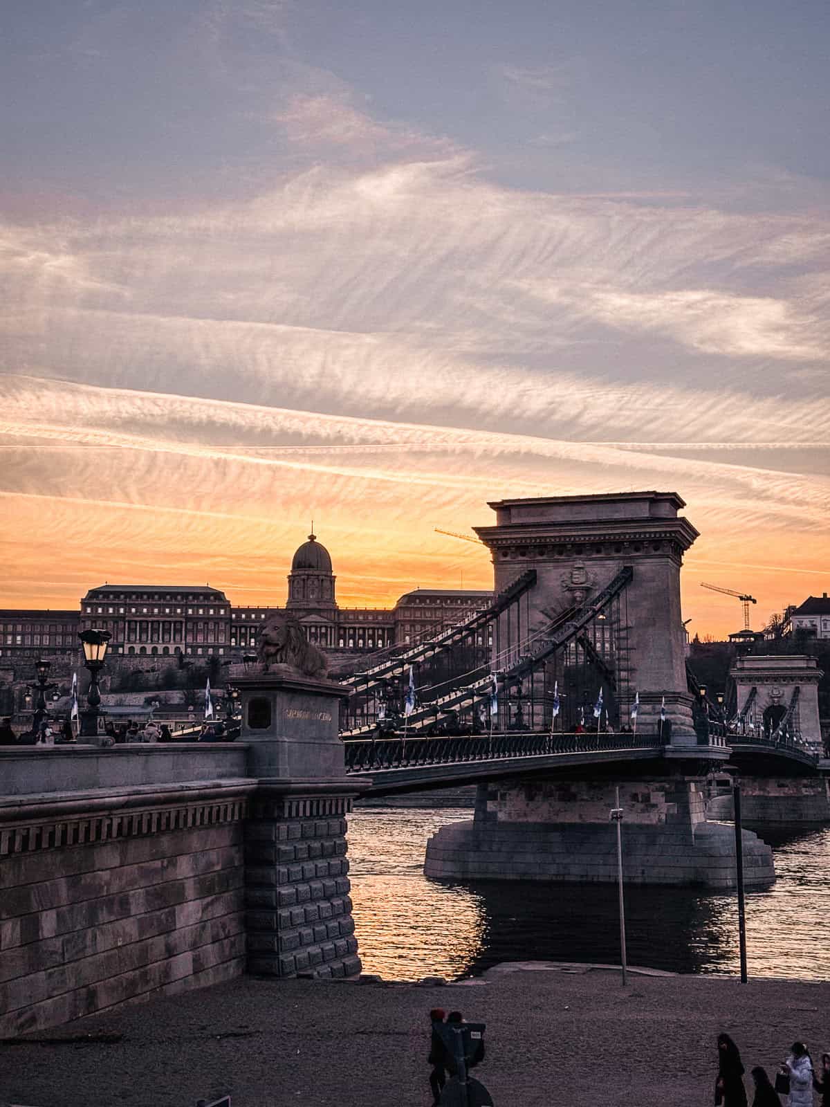 The Budapest chain bridge from a different angle, captured at sunset with an orange and pink sky, highlighting the silhouette of the bridge and historical buildings in the background.