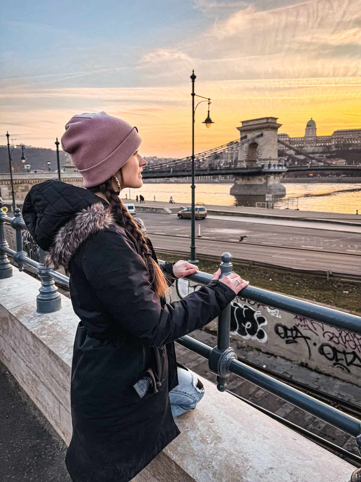 A woman in a pink beanie and black coat gazes at the Chain Bridge and historical buildings in Budapest during a beautiful sunset.