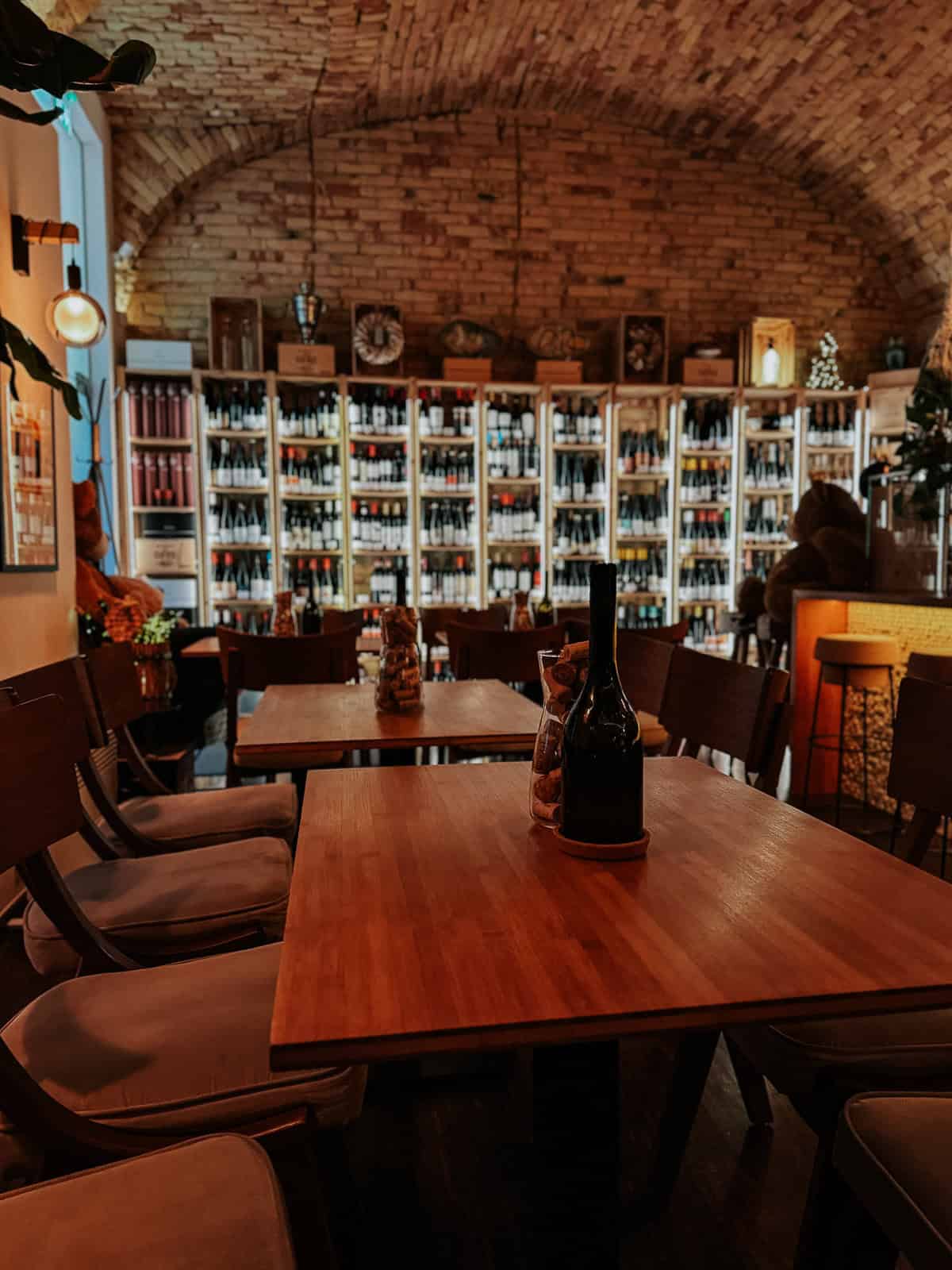 The interior of a wine bar with wooden tables, chairs, and shelves filled with wine bottles, creating a warm and inviting atmosphere.
