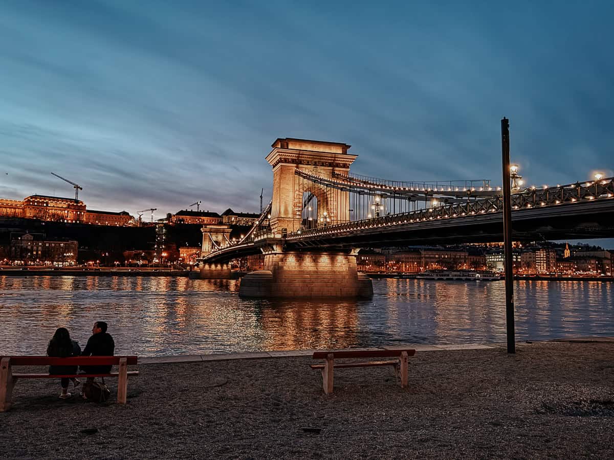 The Chain Bridge in Budapest illuminated at night, reflecting on the Danube River, with a couple sitting on a bench by the riverbank.