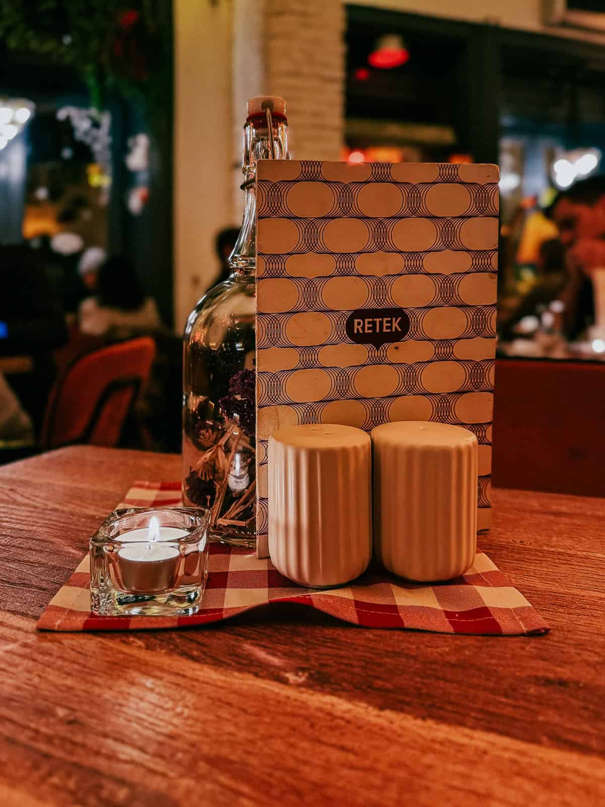 A table setting with a candle, a bottle, salt and pepper shakers, and a menu on a red-and-white checkered tablecloth.