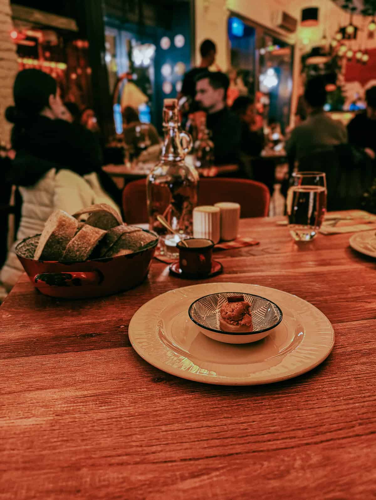 A small serving of food placed on a white plate on a wooden table, with bread in the background and a bustling restaurant scene.