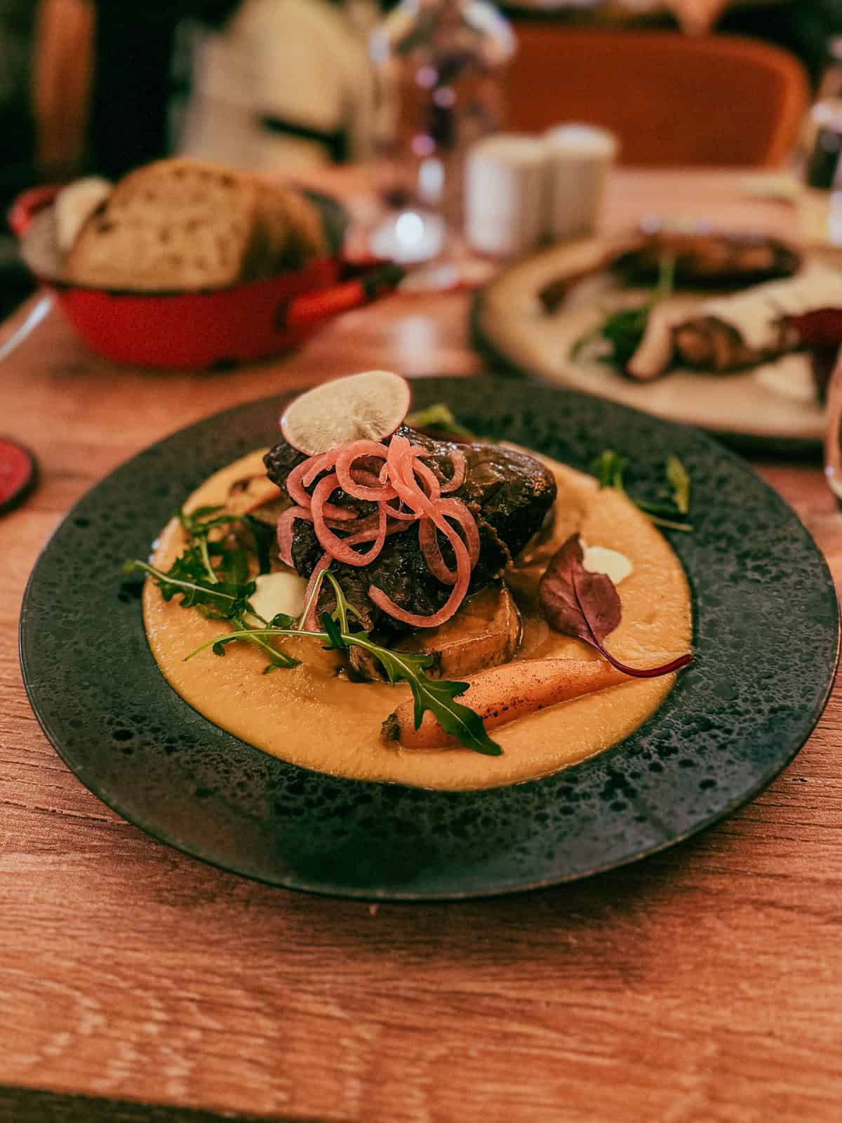 Close-up of a gourmet beef dish served on a black plate, garnished with pickled onions, greens, and a creamy sauce.