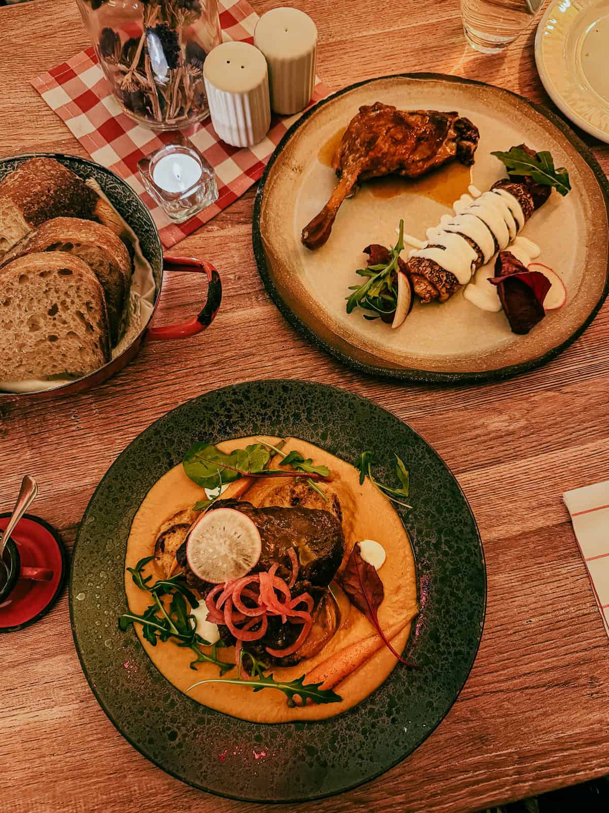 Overhead view of a wooden table with two gourmet dishes, one featuring a roasted duck leg and the other a beef dish, both accompanied by fresh garnishes.