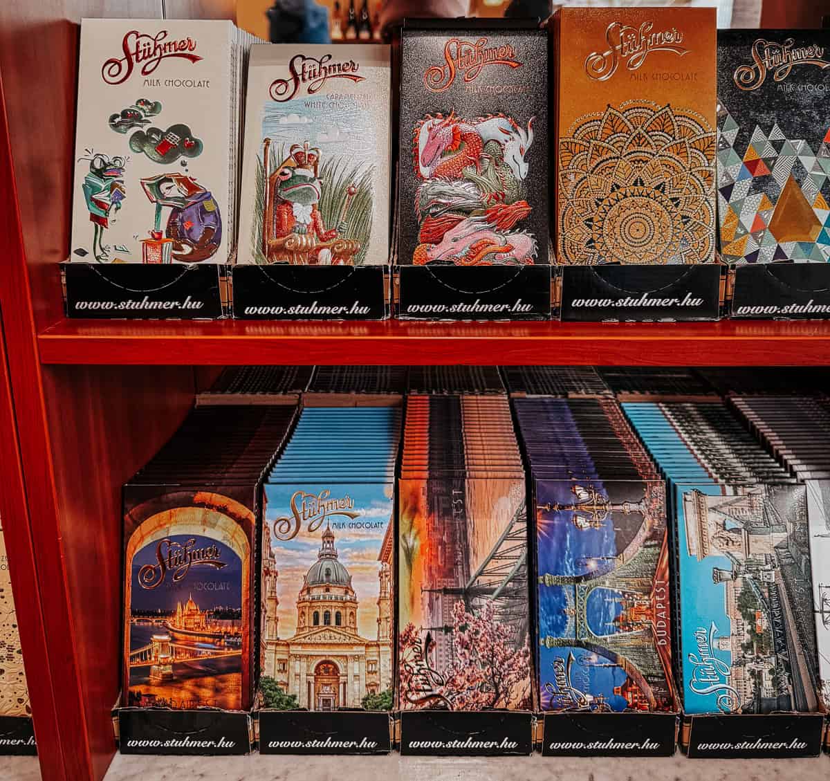 a shelf of stuhmer chocolates in budapest in various colorful packaging with iconic scenes from the city on the front