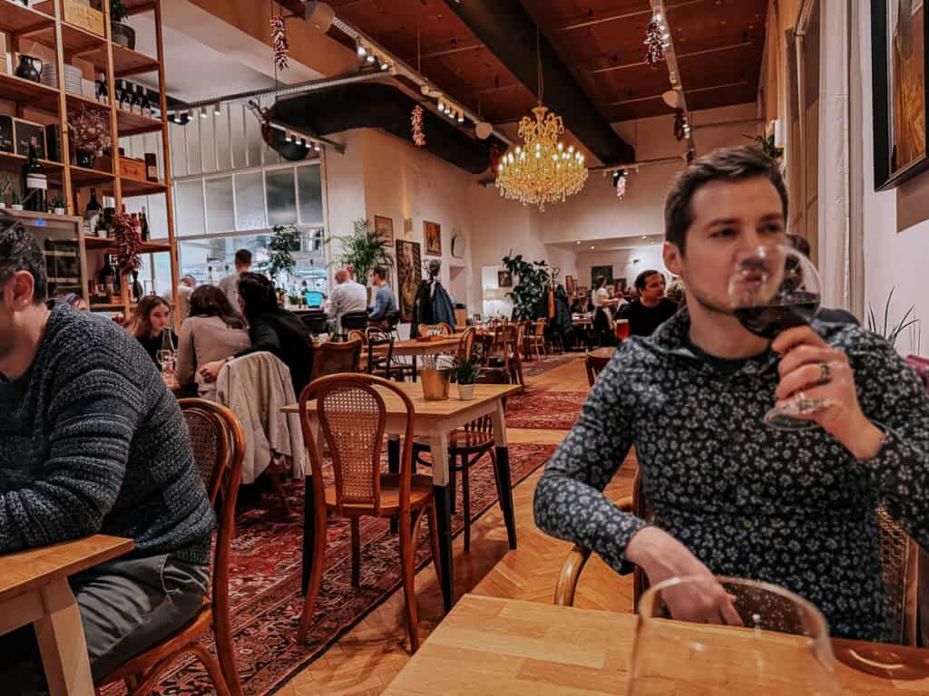 A man in a floral shirt enjoying a glass of red wine in a cozy restaurant with wooden tables and chairs. The restaurant is bustling with diners, and a chandelier hangs from the ceiling, adding to the warm ambiance.