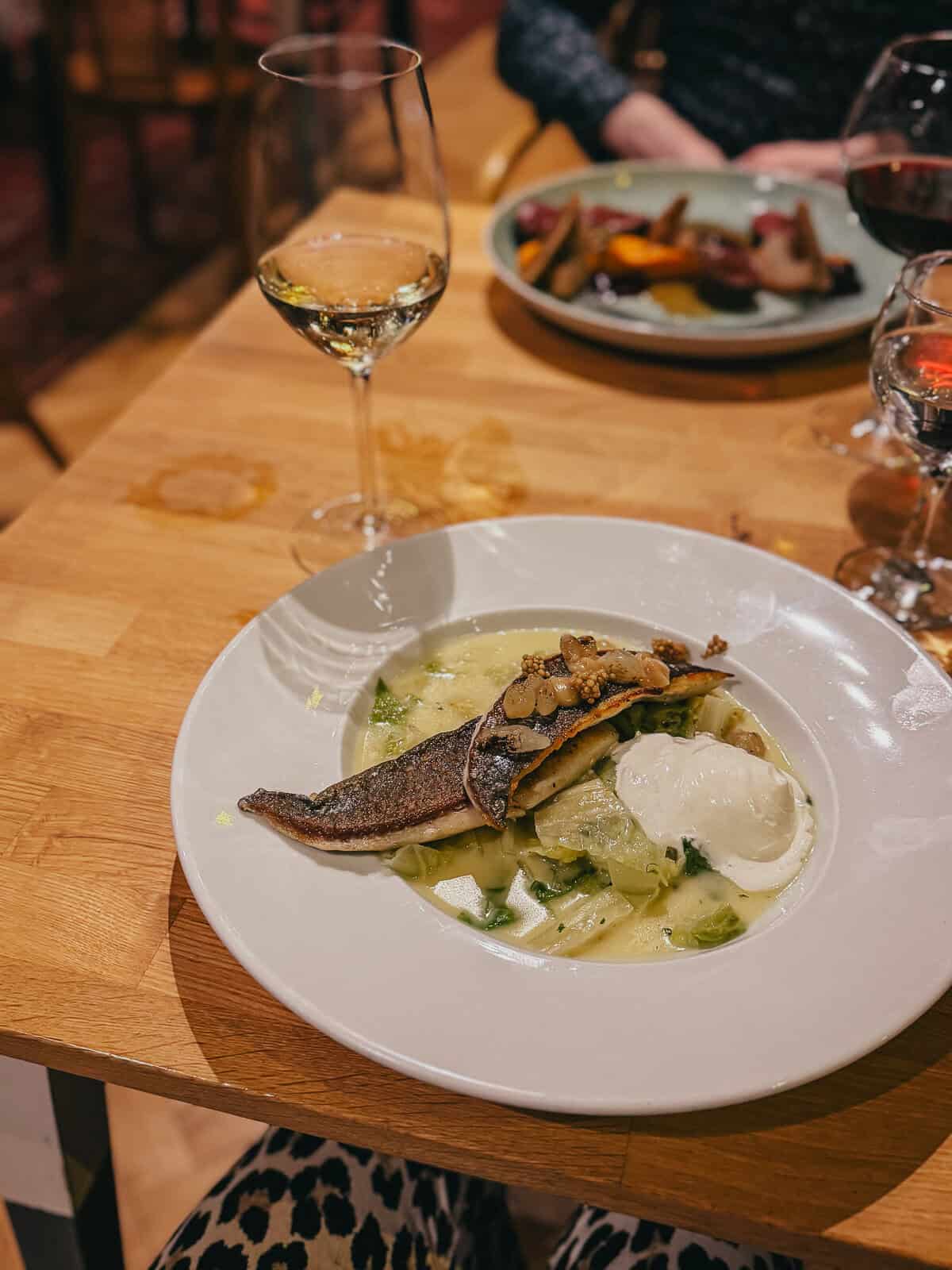 A close-up of a dish featuring a piece of fish served over a bed of vegetables and a poached egg, accompanied by a glass of white wine. The dish is artfully plated on a white plate on a wooden table.