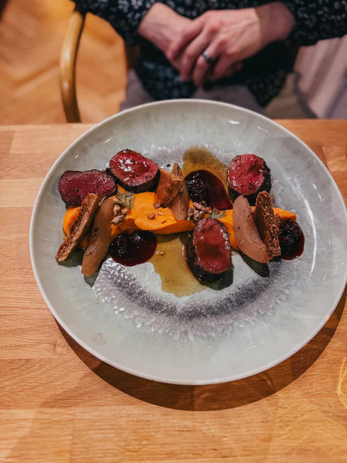 A plate of beautifully arranged food, featuring slices of meat, vegetables, and a rich sauce, on a light green ceramic plate. The dish is served on a wooden table, with someone sitting in the background.