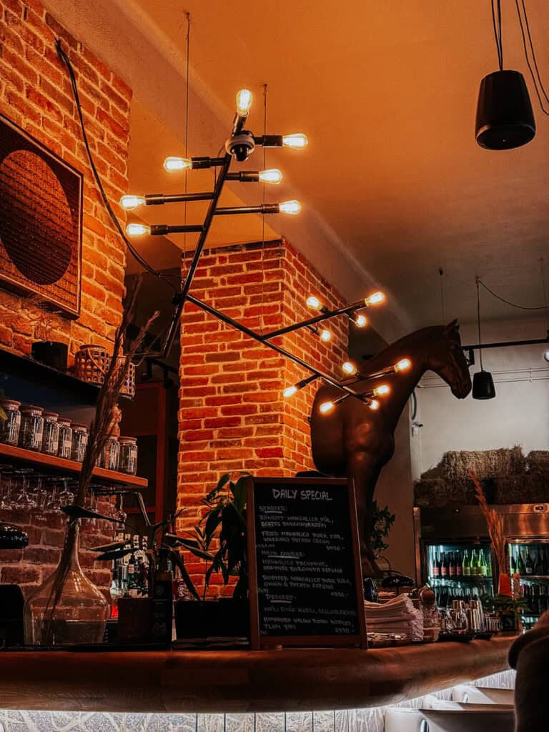 A cozy restaurant interior featuring a rustic brick wall, modern light fixtures, and a blackboard listing the daily specials. A horse statue adds a unique touch to the decor.