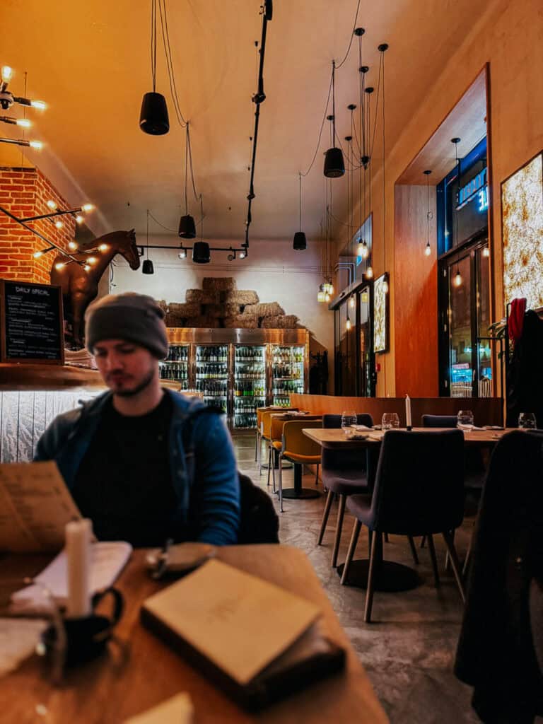 A man in a beanie sitting in a stylish restaurant with high ceilings and contemporary decor. The background shows a bar area and a refrigerator filled with bottles.