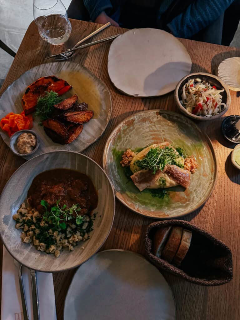 An overhead view of a table set with various dishes, including roasted vegetables, a meat stew, and a fish fillet. The table also has glasses of wine and a basket of bread.