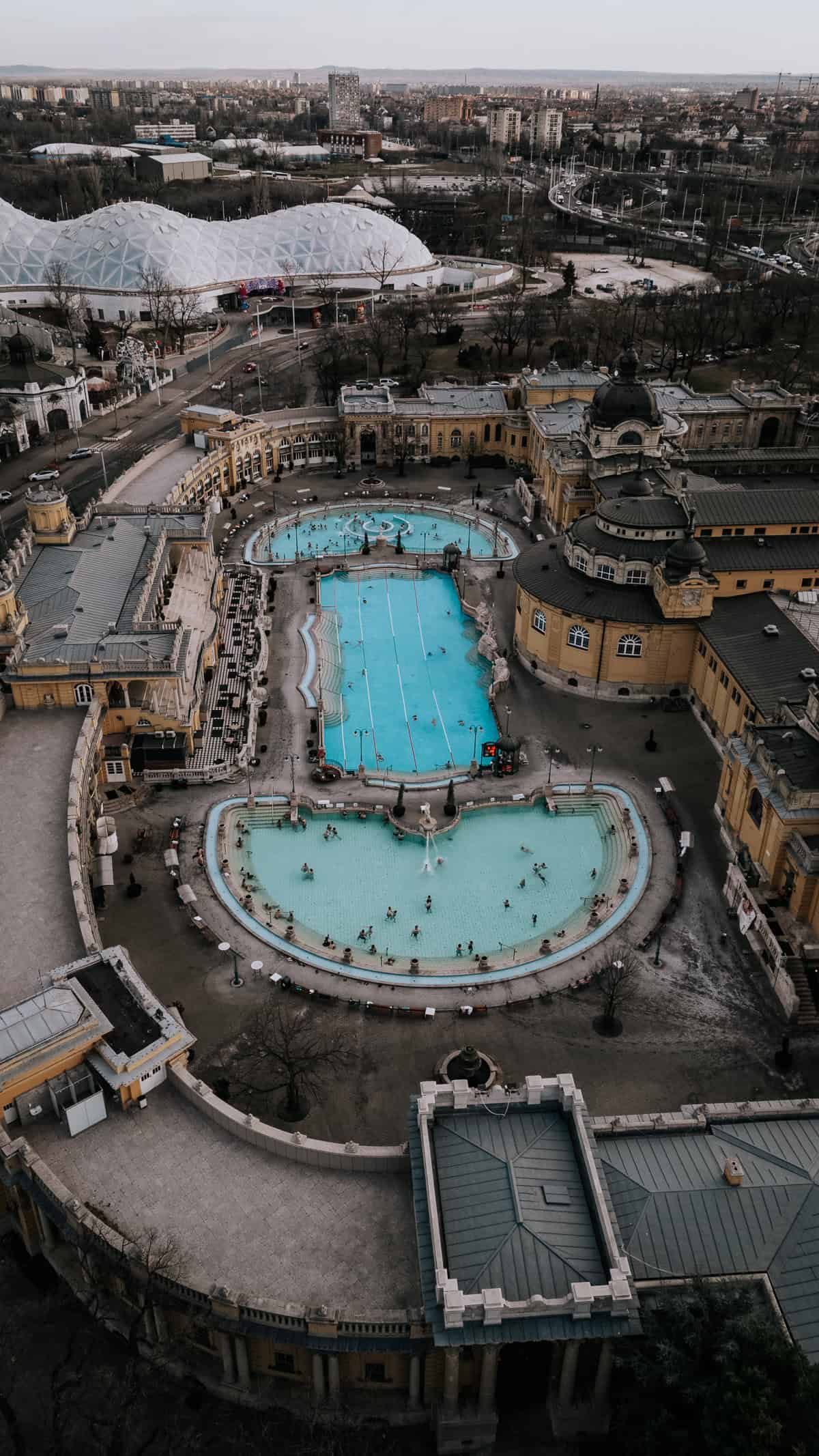An aerial view of the Széchenyi Thermal Bath in Budapest, featuring its three large outdoor pools with people swimming and relaxing, surrounded by historic yellow buildings