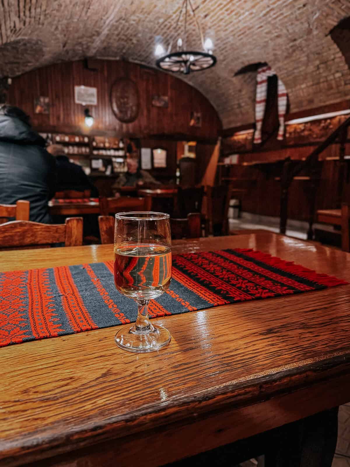 A single glass of wine on a wooden table with a red and black woven table runner in a warmly lit tavern with a stone ceiling and wooden decor.