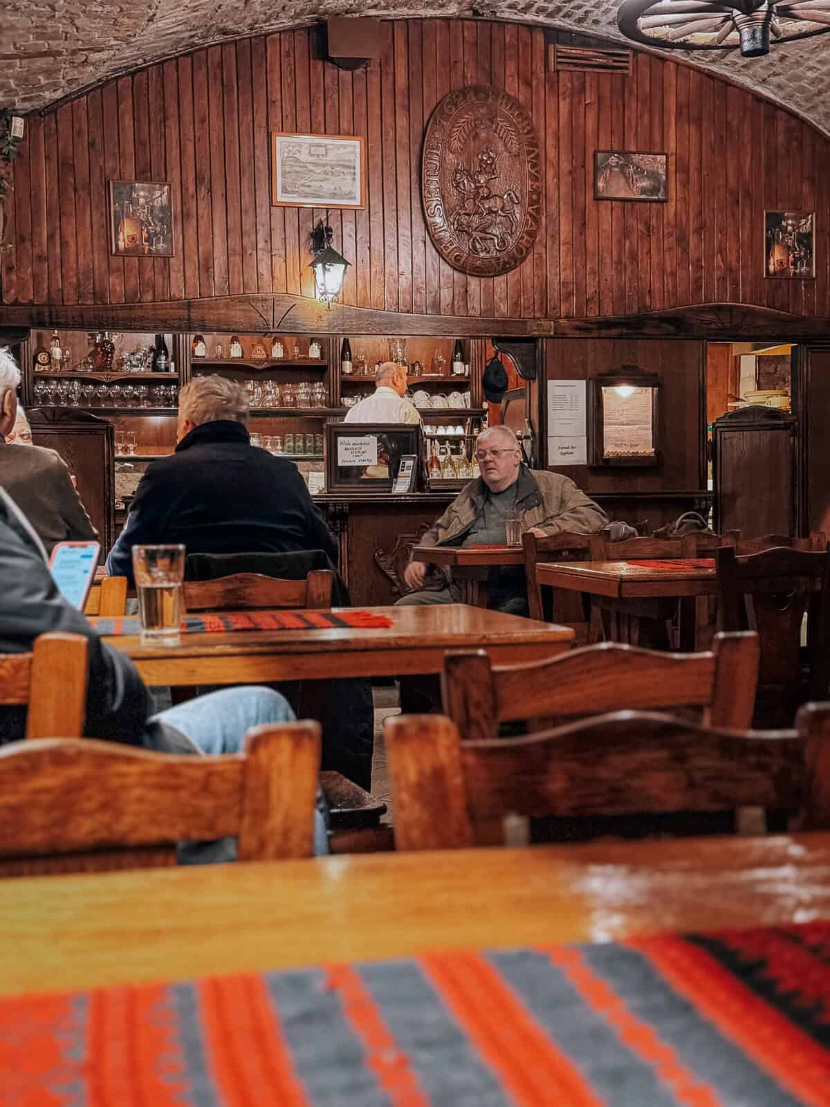 An interior view of a rustic tavern with wooden walls and ceiling, a bar area with various bottles and glasses, and patrons seated at wooden tables, engaging in conversation.