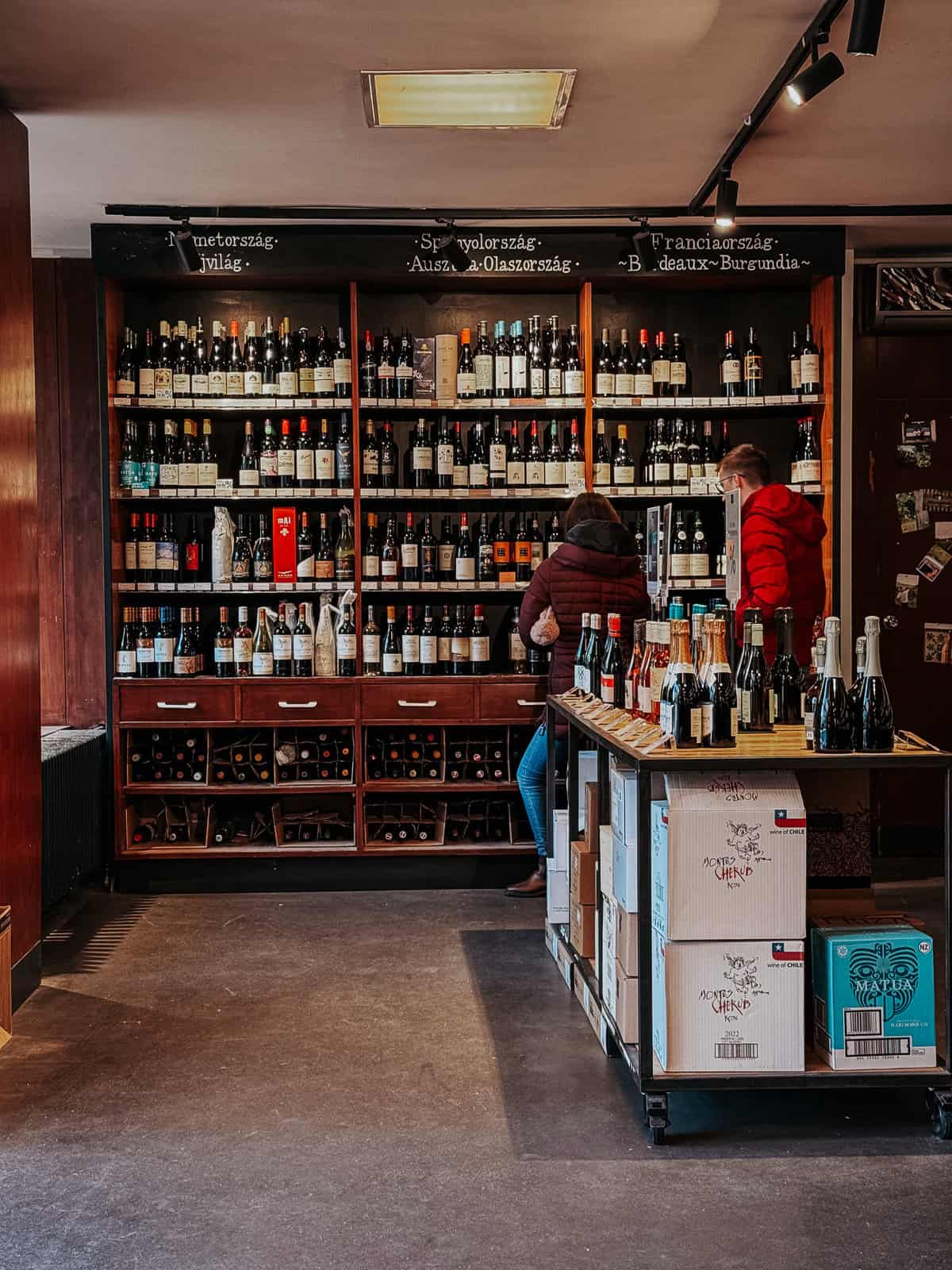 A well-stocked wine shop with shelves filled with various wine bottles. Two customers are browsing the selection, and boxes of wine are stacked in the foreground.