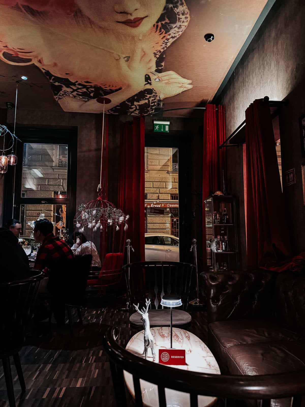 The interior of a chic bar with moody lighting, featuring a wall mural of a woman's face, plush seating, and patrons enjoying their drinks. The decor includes birdcage-style light fixtures and red curtains.