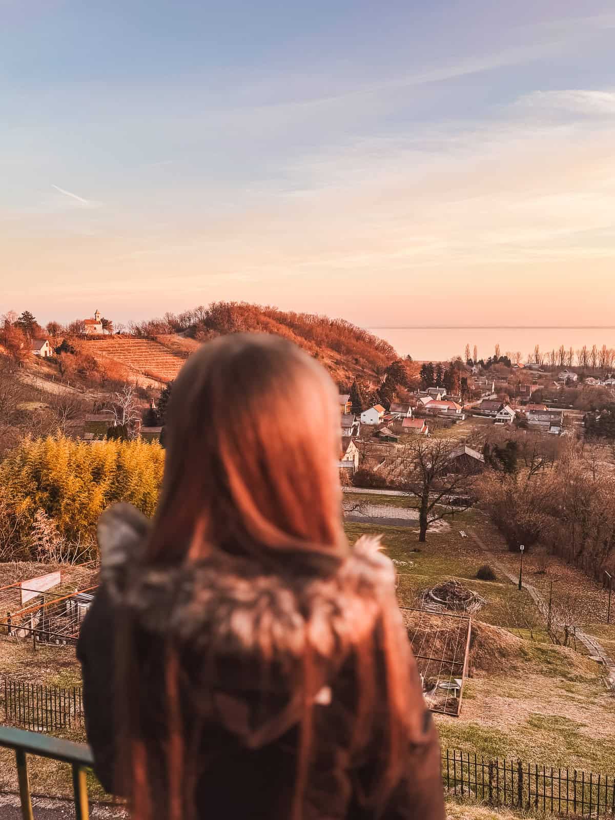 A woman with long hair and a fur-collared coat stands on a balcony overlooking a picturesque village and rolling hills at sunset. The sky is a gradient of warm colors, adding a serene and beautiful backdrop.