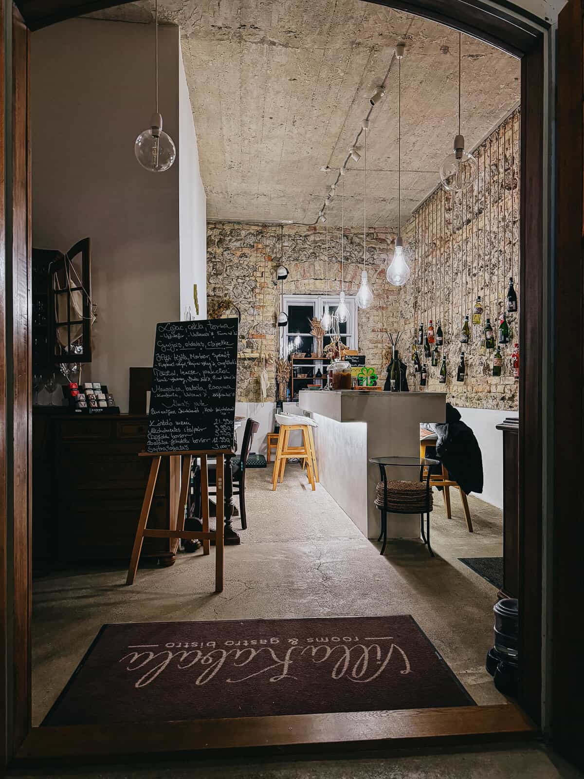 An inviting interior of a wine bar or bistro with a mix of modern and rustic decor. The space features a bar area, stools, a chalkboard menu, and ambient lighting, creating a cozy and welcoming atmosphere.