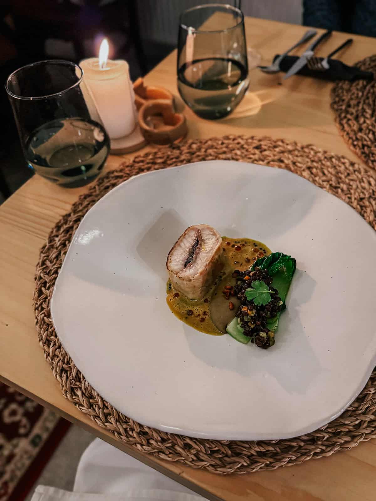 A beautifully plated dish featuring a piece of fish atop a yellow sauce, with greens and grains on the side. The setting includes two glasses of water and a lit candle, creating a cozy dining atmosphere.