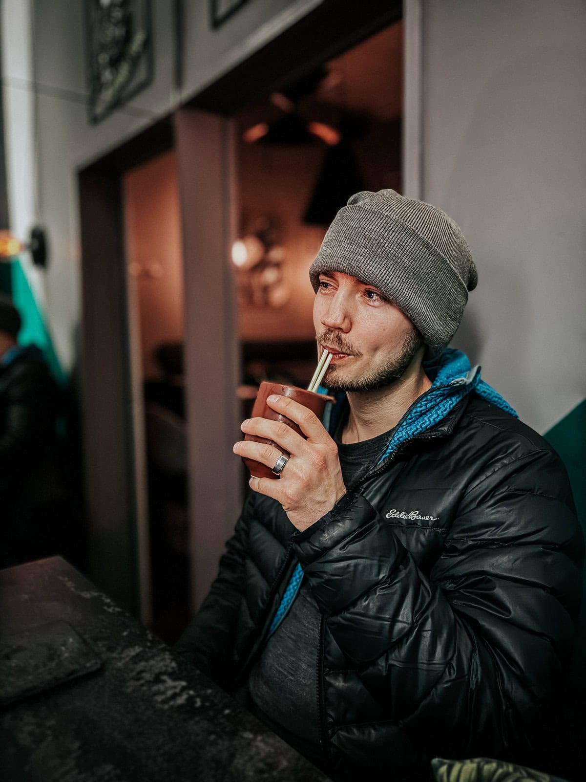 A man in a winter jacket and beanie enjoying a drink with a straw at a cozy bar. The background is dimly lit, creating a warm atmosphere.