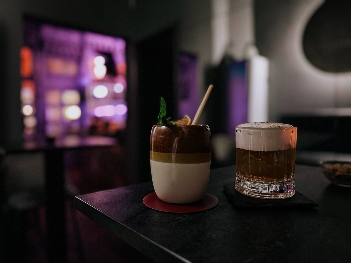 Two cocktails on a bar counter, one topped with foam and the other garnished with mint and citrus. The dim lighting and blurred background give the bar a sophisticated ambiance.