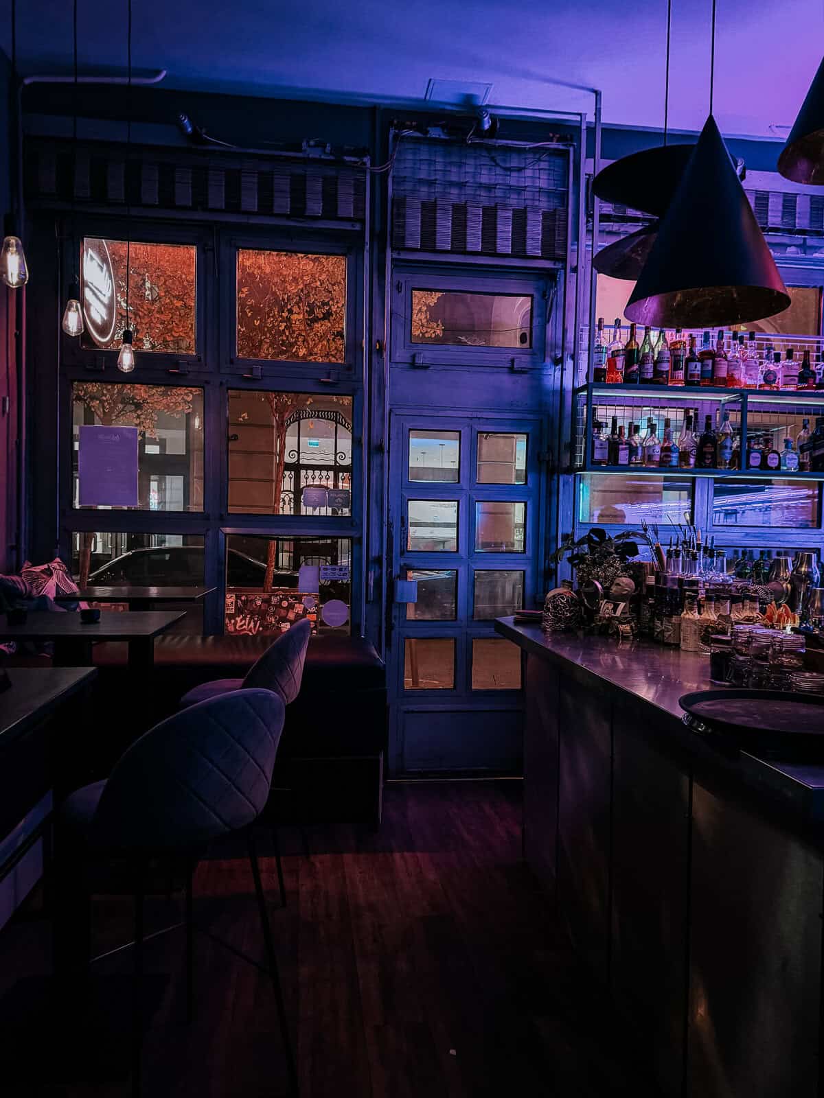 The interior of a trendy bar with a blue and purple neon glow, featuring a well-stocked bar, high chairs, and large windows. The decor is modern and inviting.