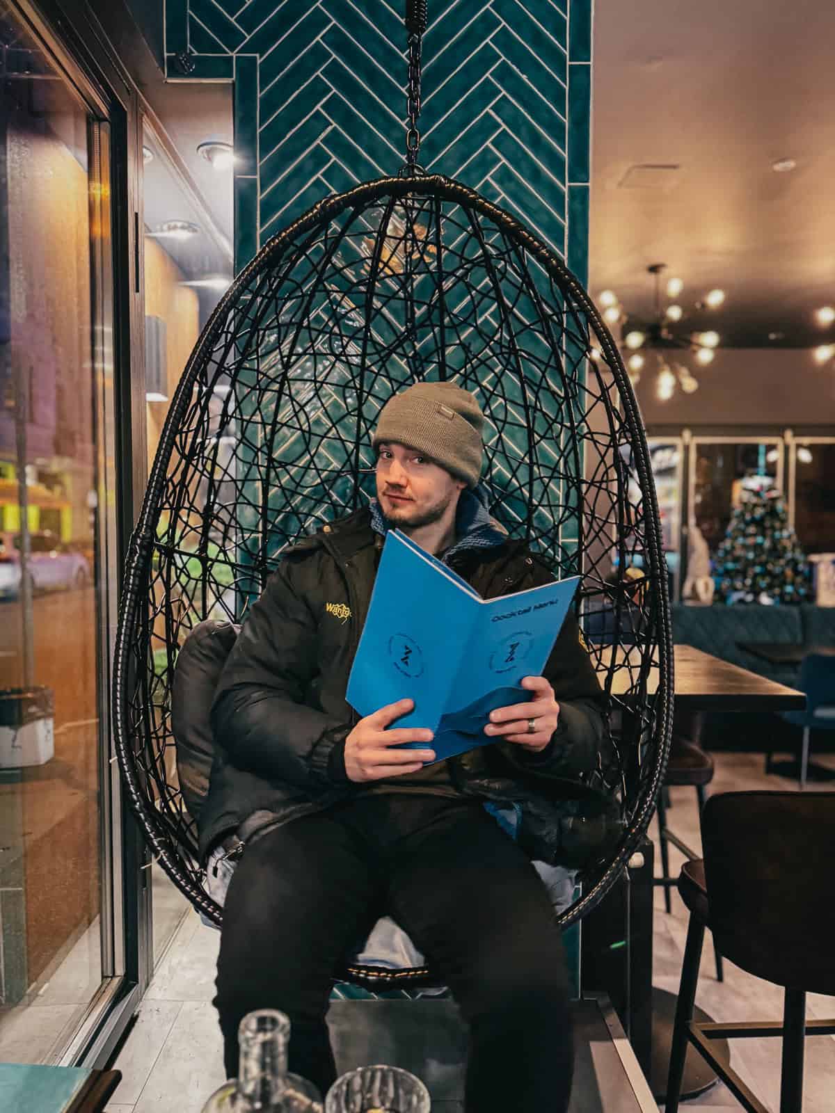 A person sitting in a hanging wicker chair inside a bar, reading a blue menu, with a green tiled wall behind.