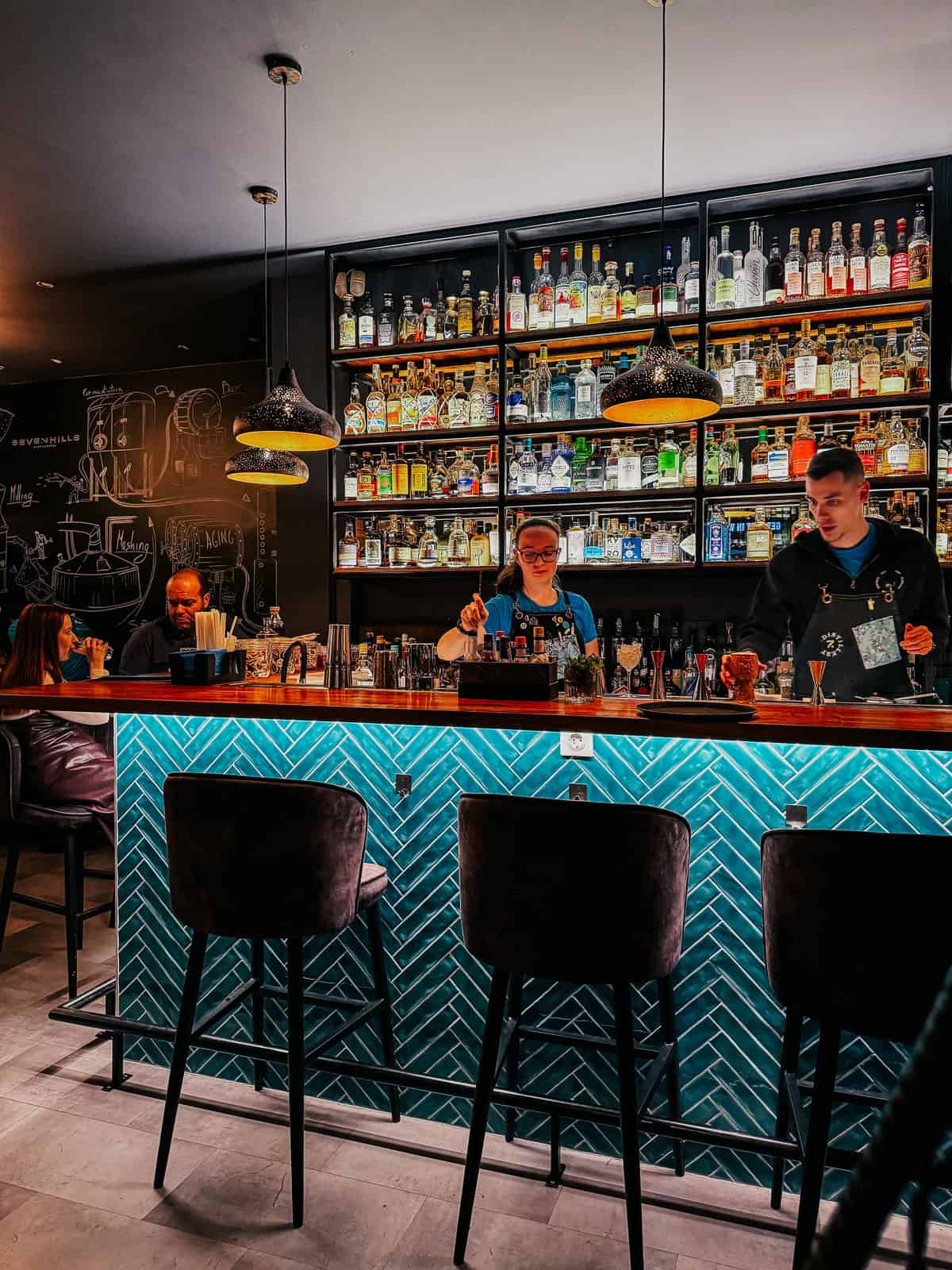 A brightly lit bar with a teal herringbone tile design, shelves filled with various spirits, and bartenders preparing drinks.