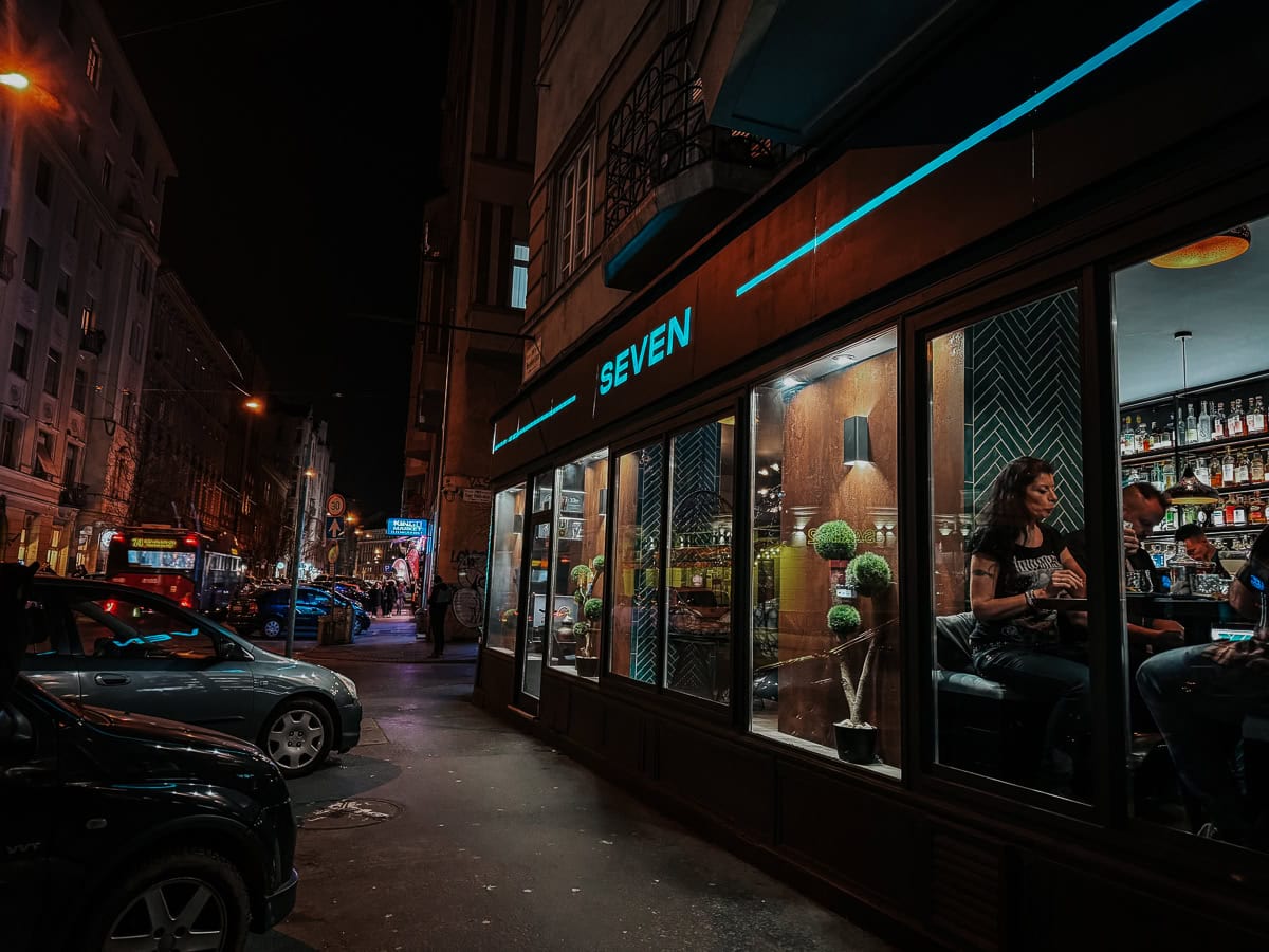 A nighttime street view of the bar 'Seven,' showing people inside enjoying drinks, with the bar’s neon sign glowing