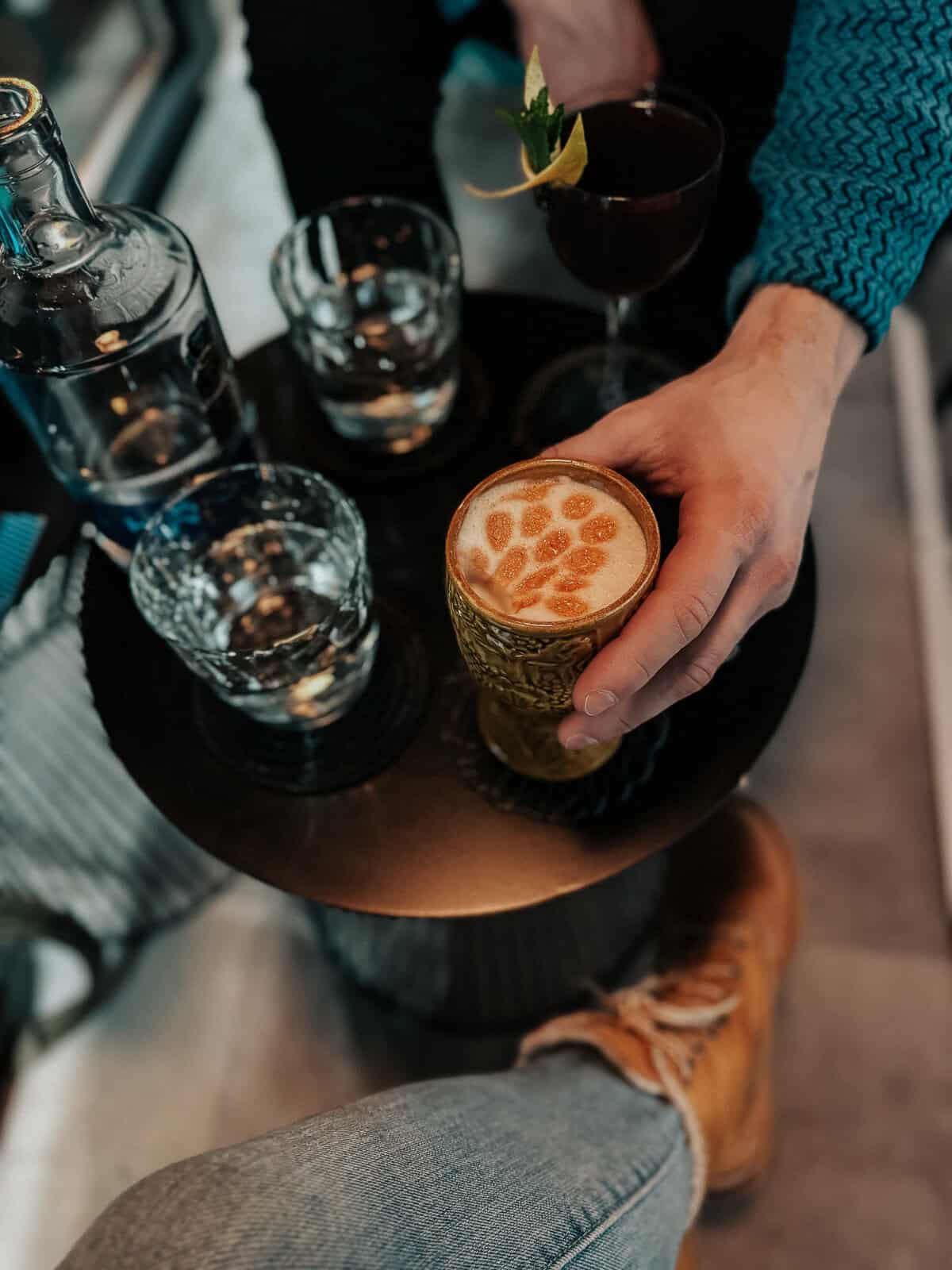 Close-up of a person’s hand holding a garnished cocktail, with additional drinks and a water bottle on a small table.