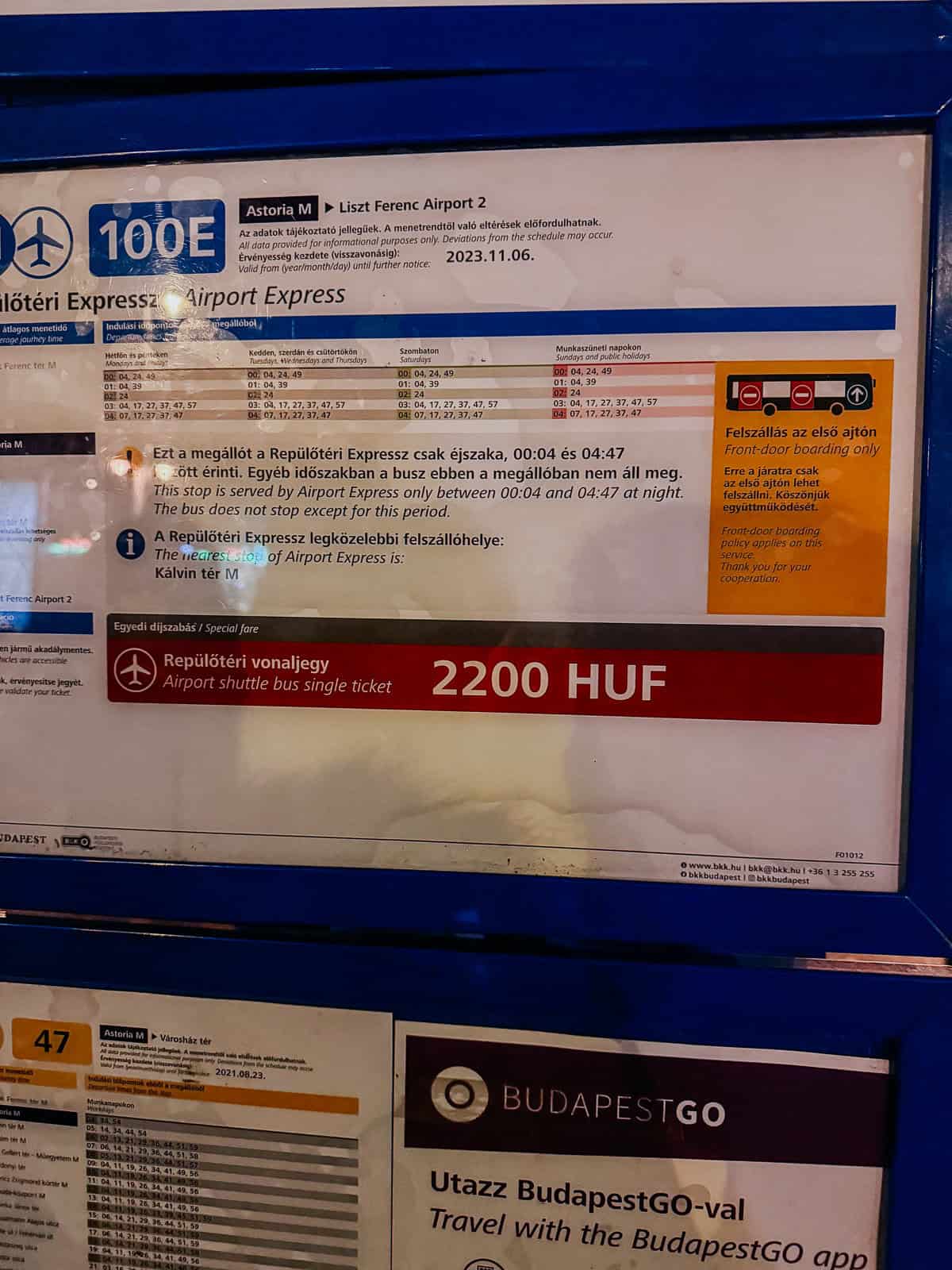 A sign for the 100E Airport Express bus at a bus stop in Budapest, displaying the schedule, fare of 2200 HUF, and instructions for front-door boarding only.