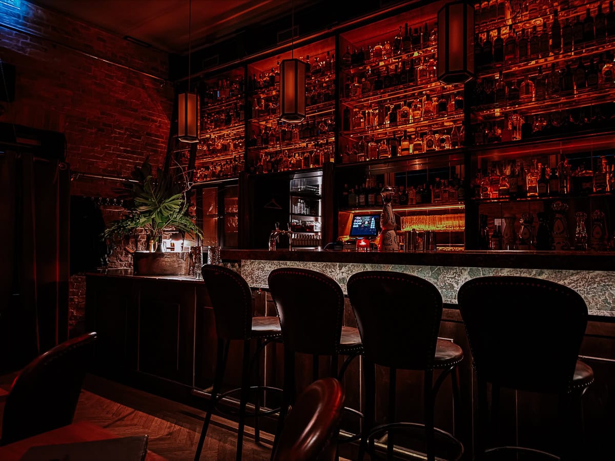 A dimly lit bar with dark wooden furniture and shelves filled with various spirits, creating a warm and intimate atmosphere.